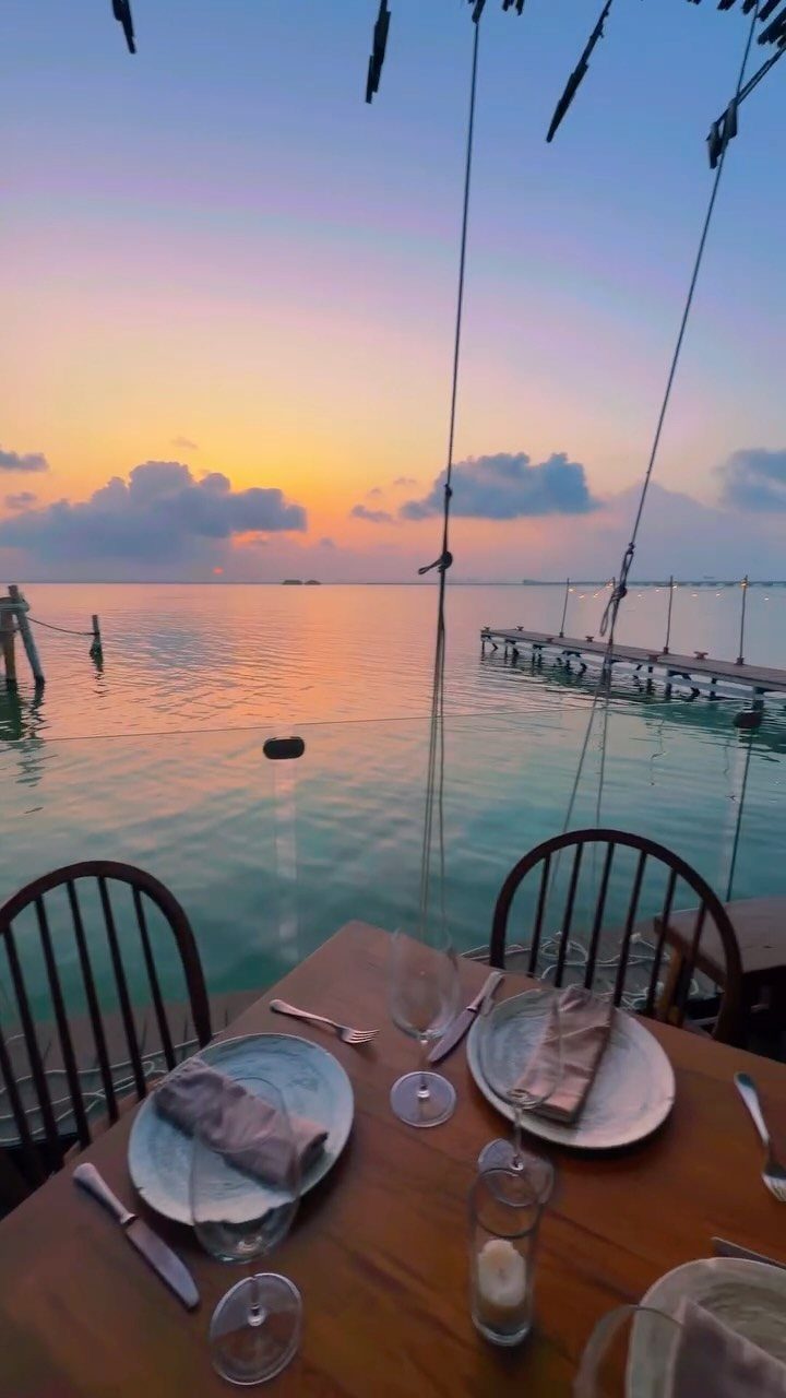 Romantic dinner table set on a deck overlooking a sunset ocean beach scene.
