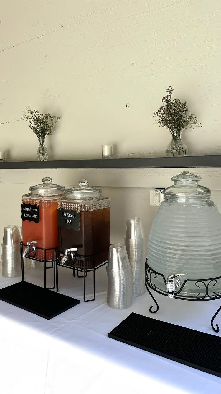 Three glass beverage dispensers with different drinks on a white table with silver cups.