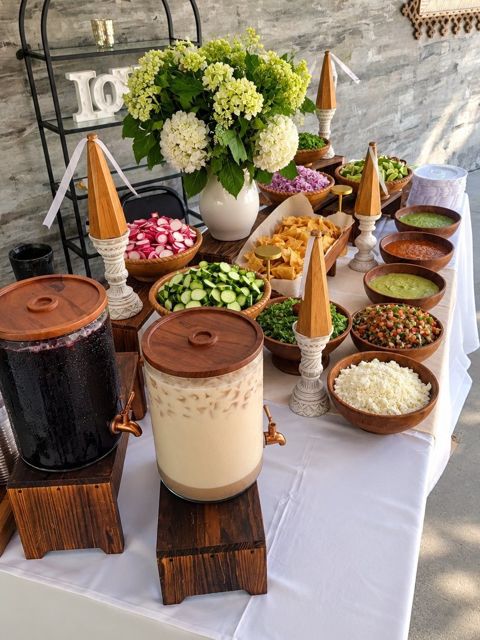 Taco station with bowls of fresh toppings and beverage dispensers on white tablecloth.