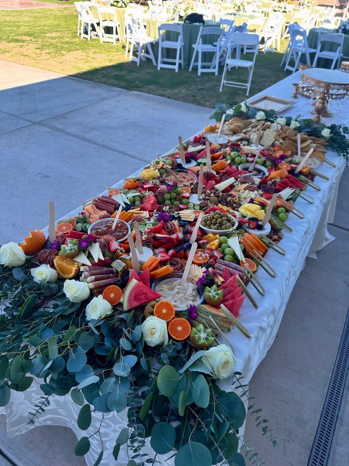 Grazing table with assorted fruits, cheeses, and meats decorated with white roses and eucalyptus.