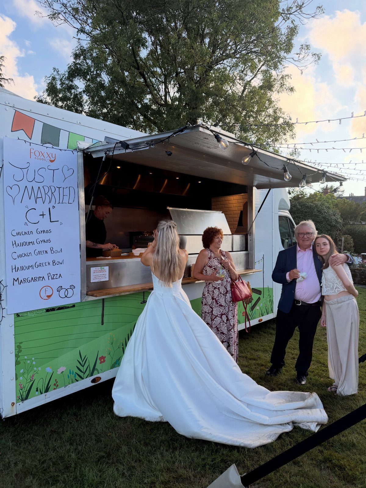 Bride and guests ordering food from a decorated wedding food truck outdoors.