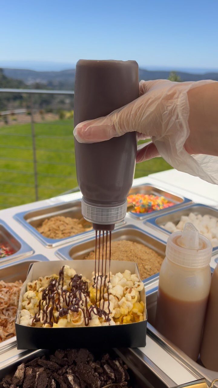 Chocolate sauce being drizzled over popcorn at a customized snack station outdoors.