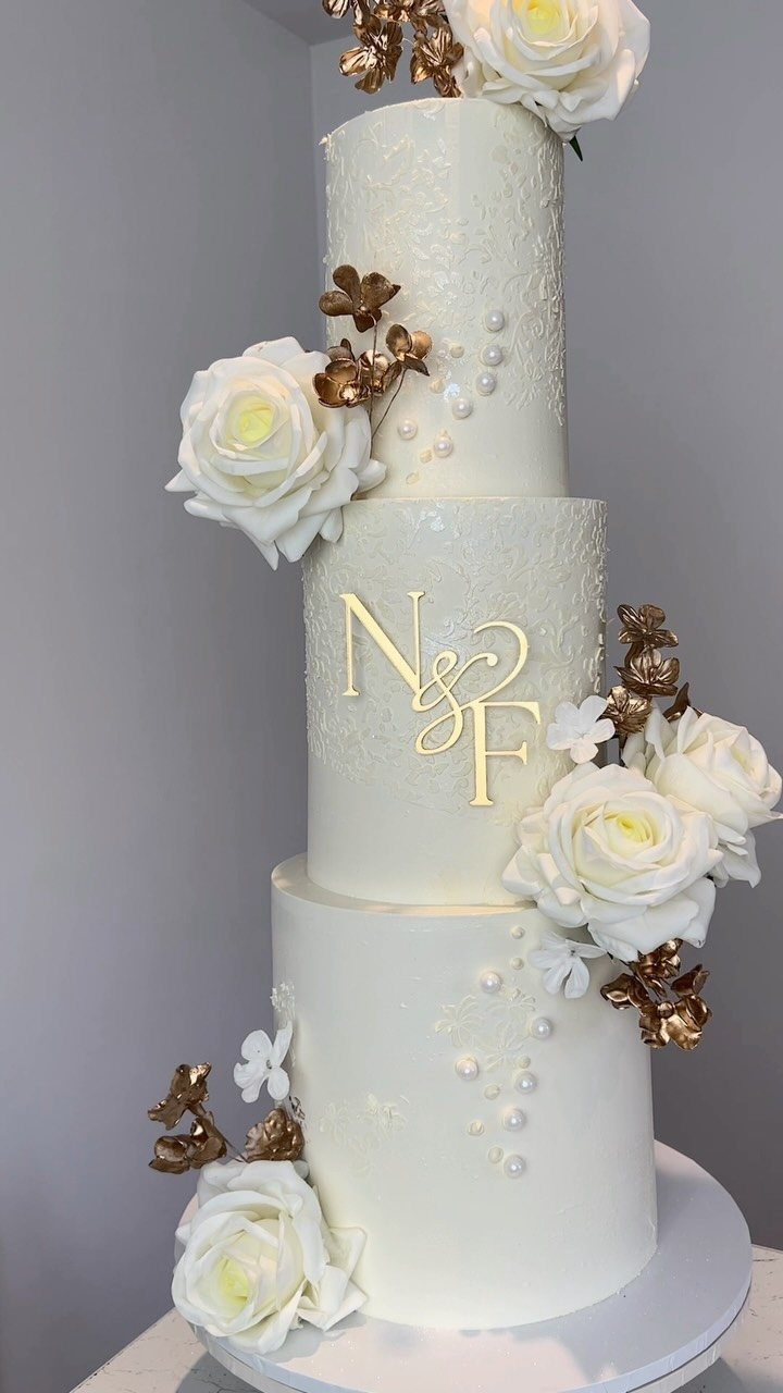Three-tier white wedding cake with gold initials, white roses, and gold leaf accents.