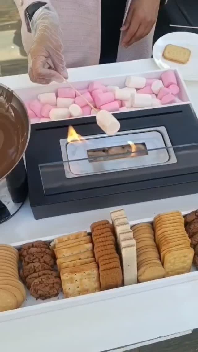 Hand roasting a marshmallow over a tabletop fire pit with crackers and marshmallows.
