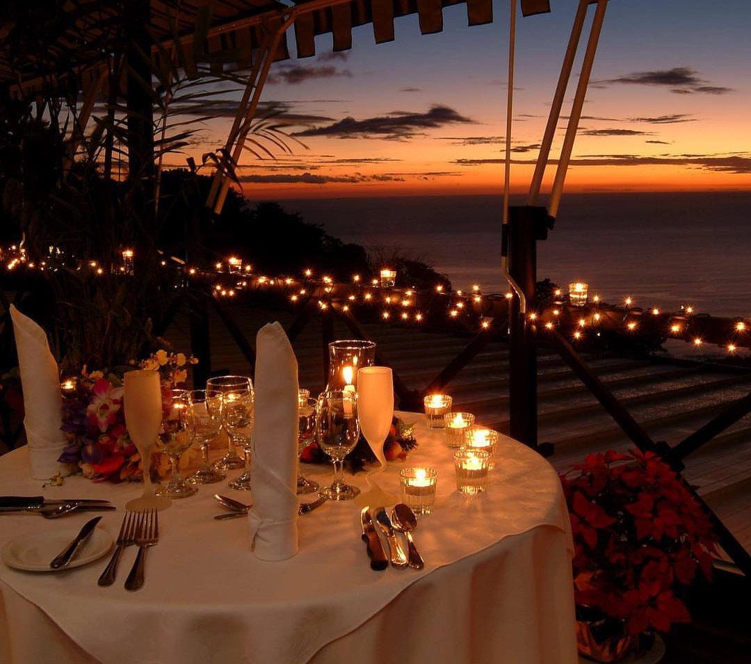 Romantic dinner table set with candles and glassware overlooking an ocean sunset.