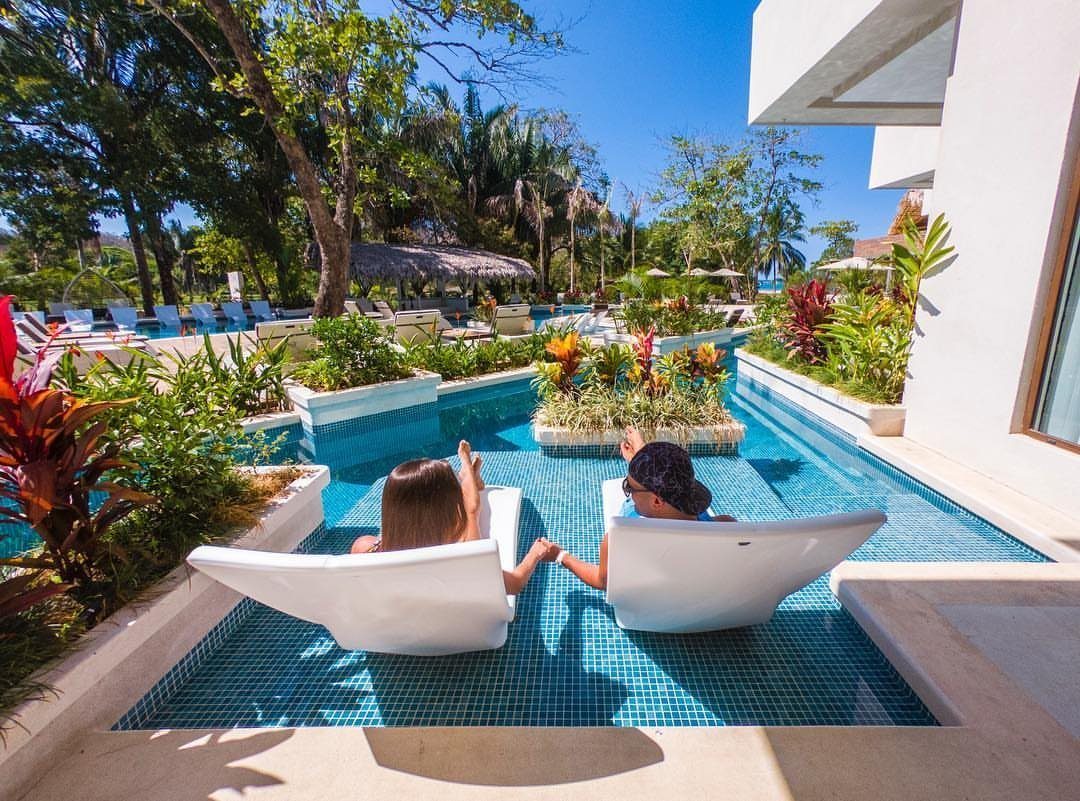Couple holding hands while relaxing in white lounge chairs inside a tropical pool.