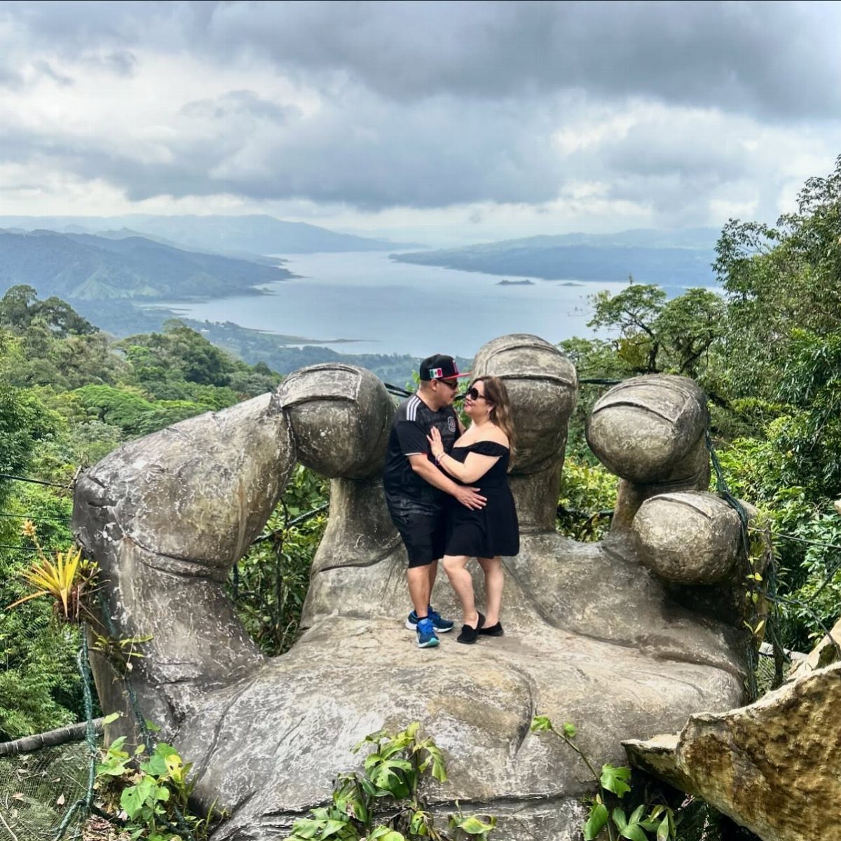 Couple kissing on a giant stone hand sculpture overlooking a lake and mountains.