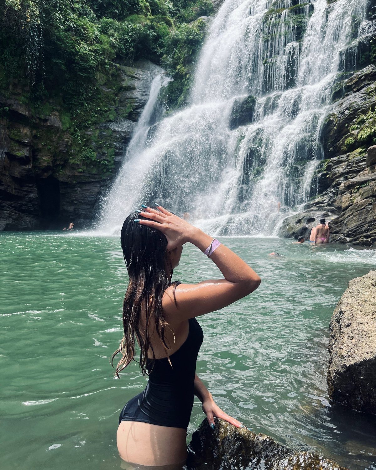 Woman in black swimsuit standing in turquoise water before a tropical waterfall