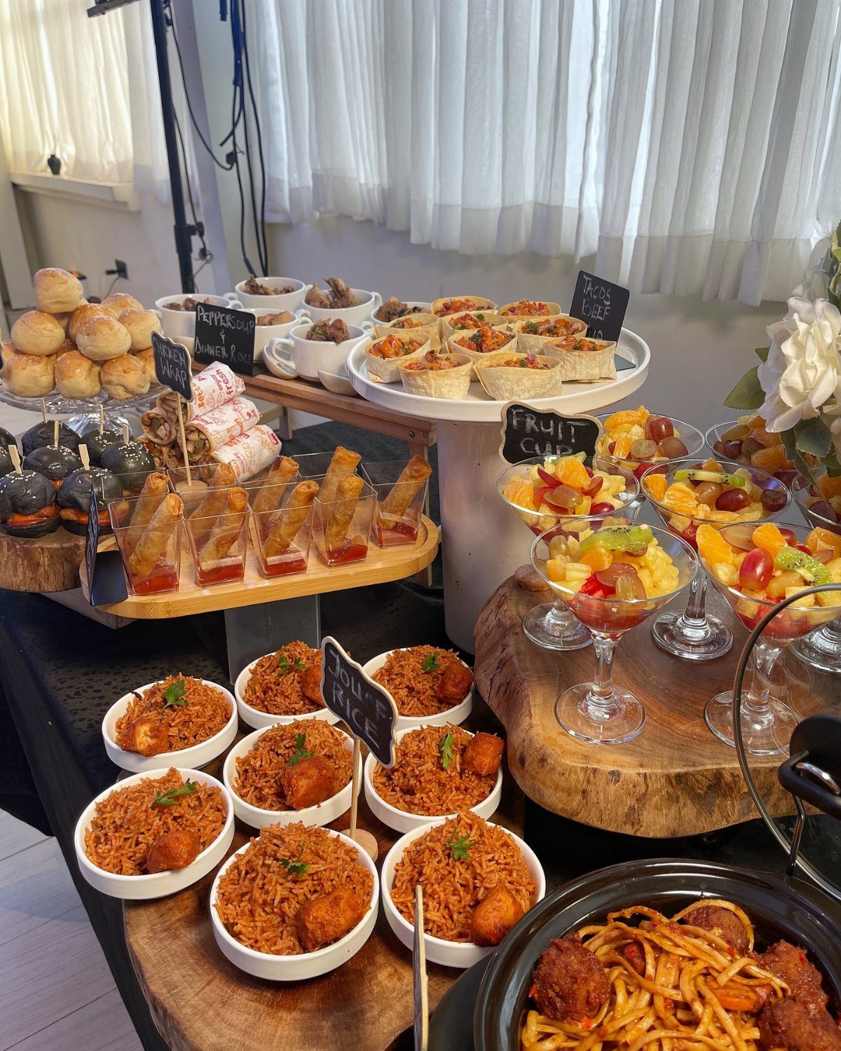 Buffet spread with jollof rice, fruit salads, and appetizers on wooden boards.