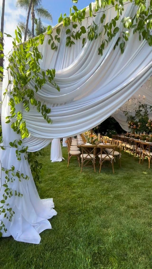 White draped fabric canopy with green vines over wooden tables on green grass.