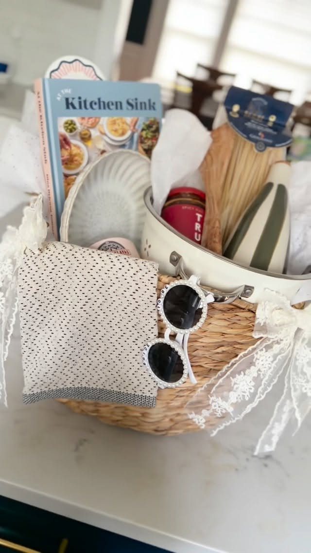 Wicker gift basket containing a cookbook, kitchenware, and a patterned towel on marble.