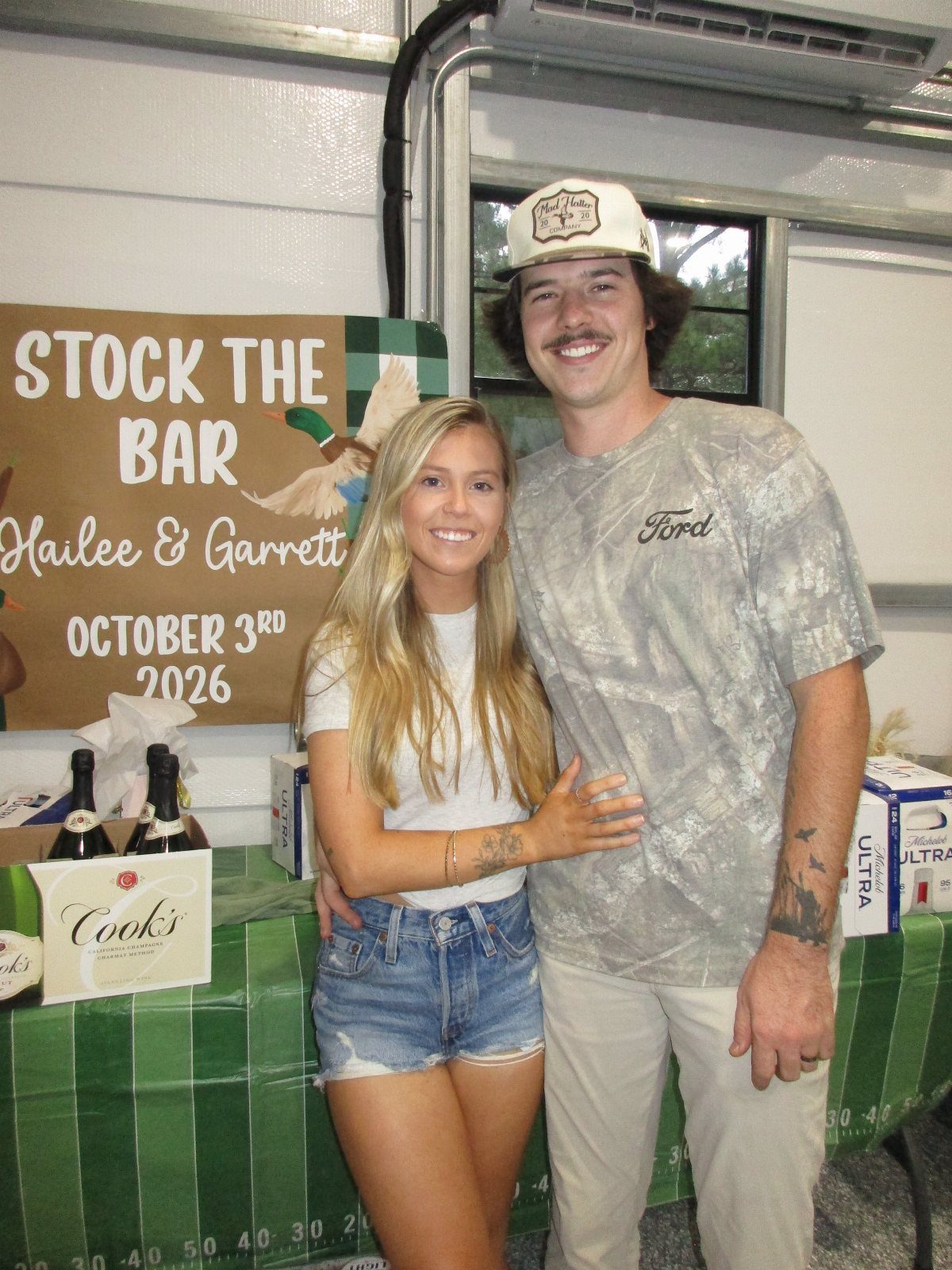 Couple posing in front of a Stock the Bar sign with alcohol gifts.