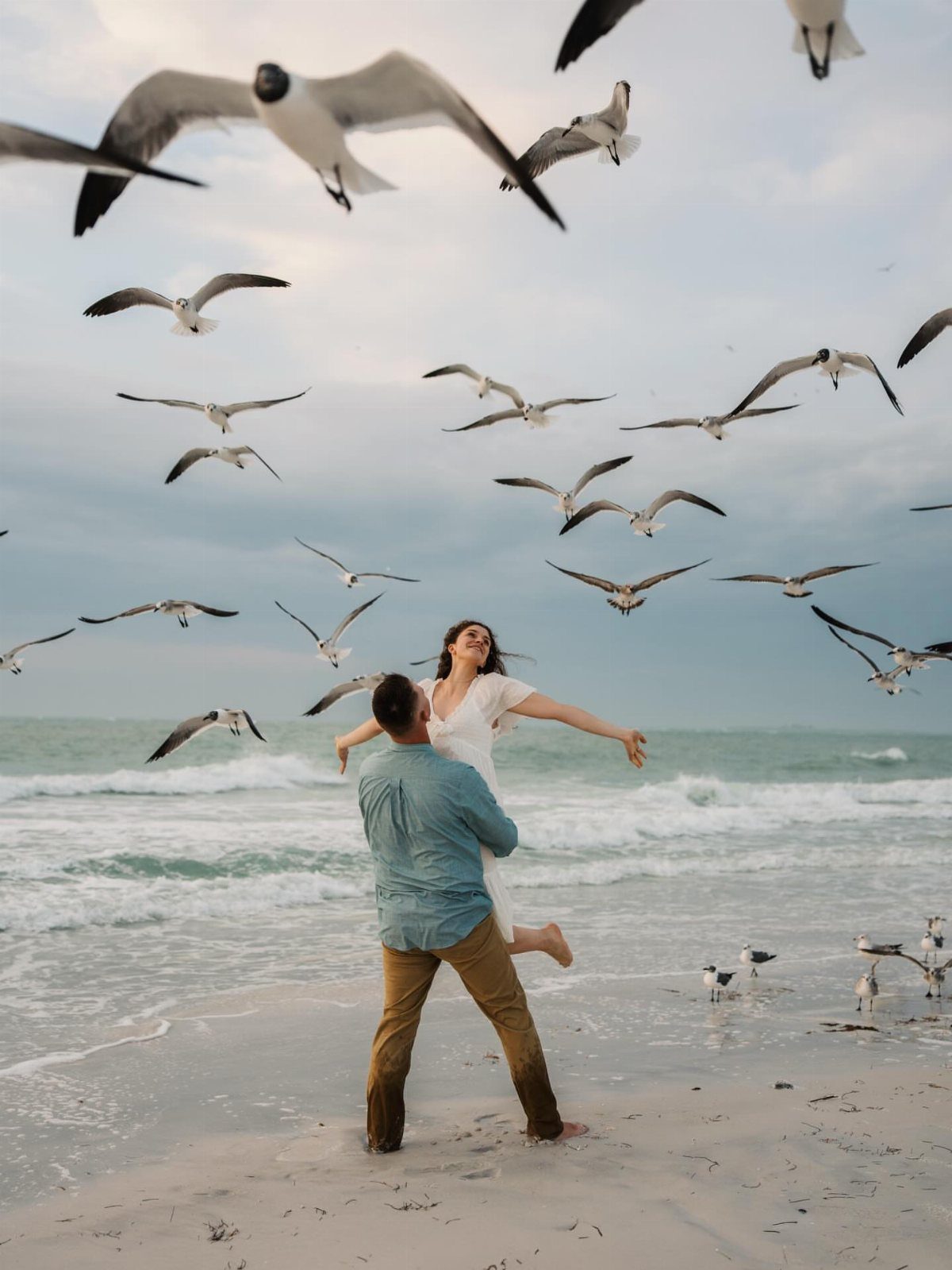 Couple embracing on a sandy beach surrounded by many flying seagulls at dusk.