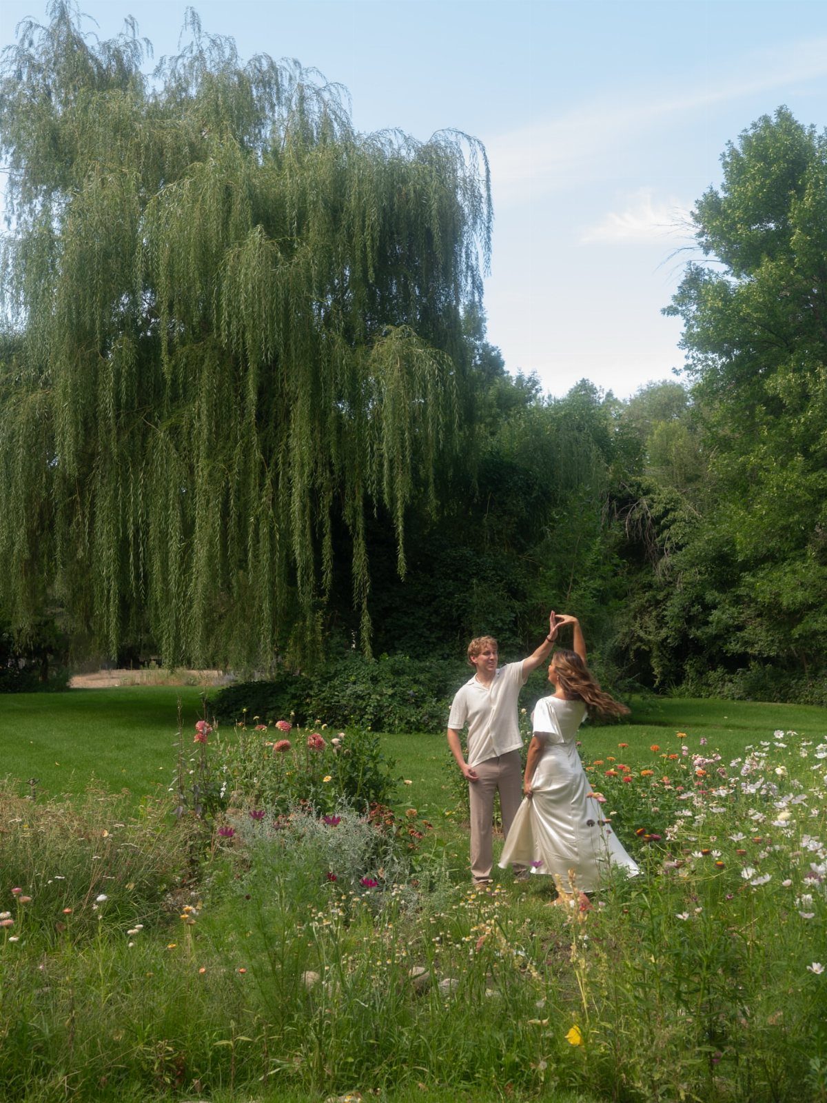 Couple in white dancing in a wildflower meadow with a weeping willow tree.