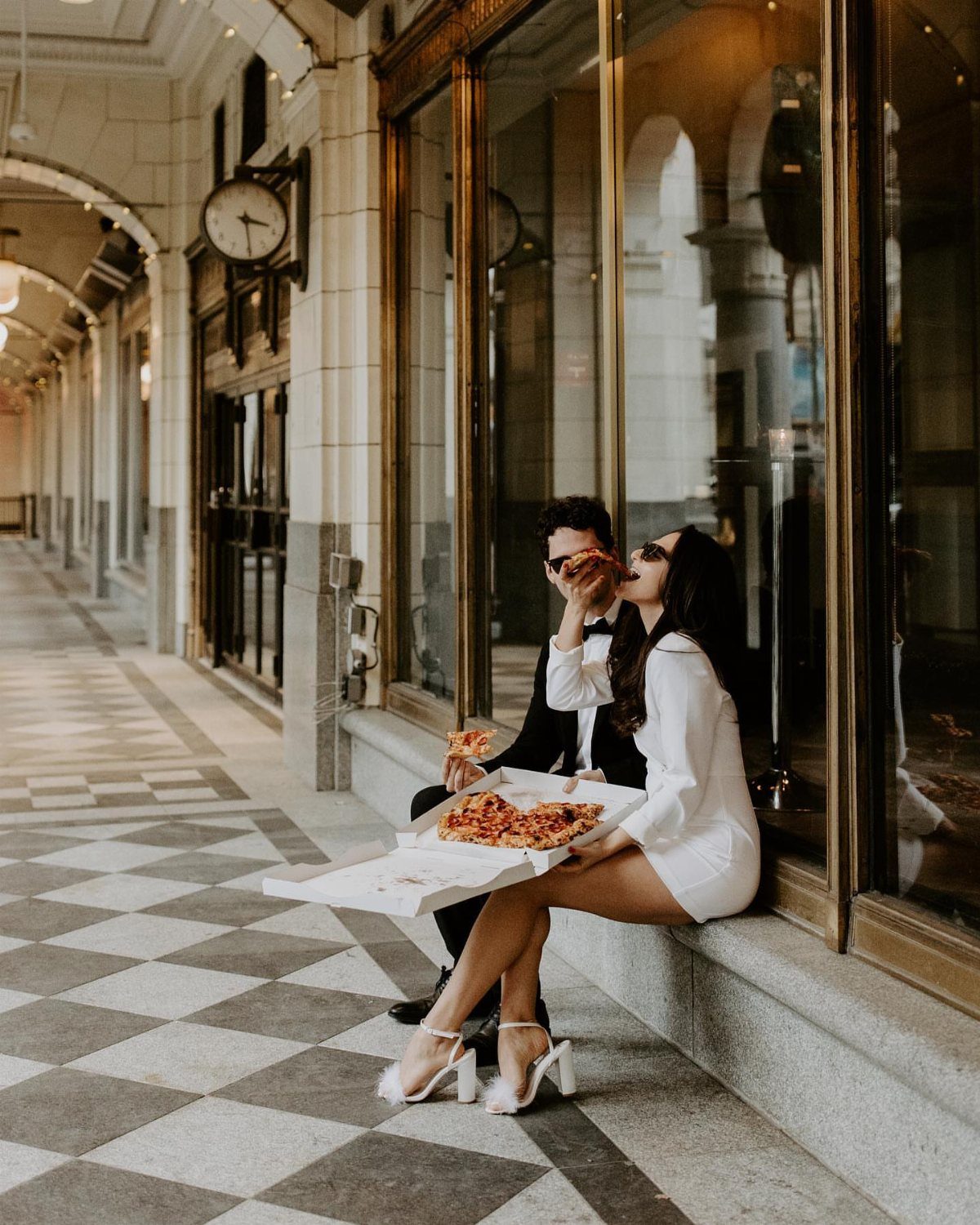 Couple in formal attire eating pizza while sitting outside a city storefront