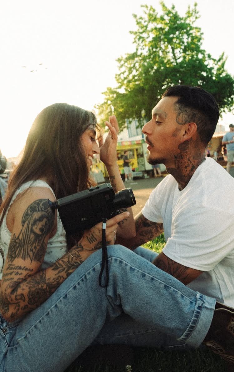 Tattooed couple sitting on grass, woman holding a vintage camera while smiling.