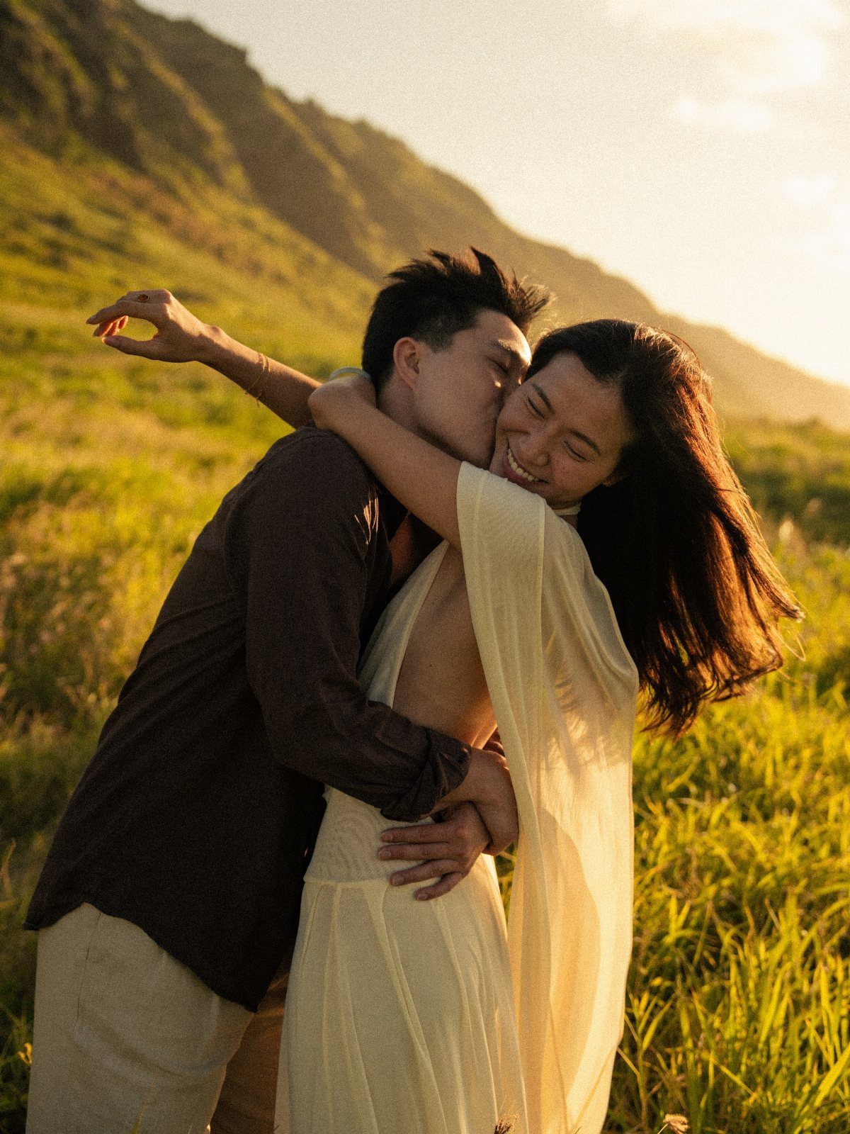Couple embracing in a golden grassy field during a warm sunset with mountains.