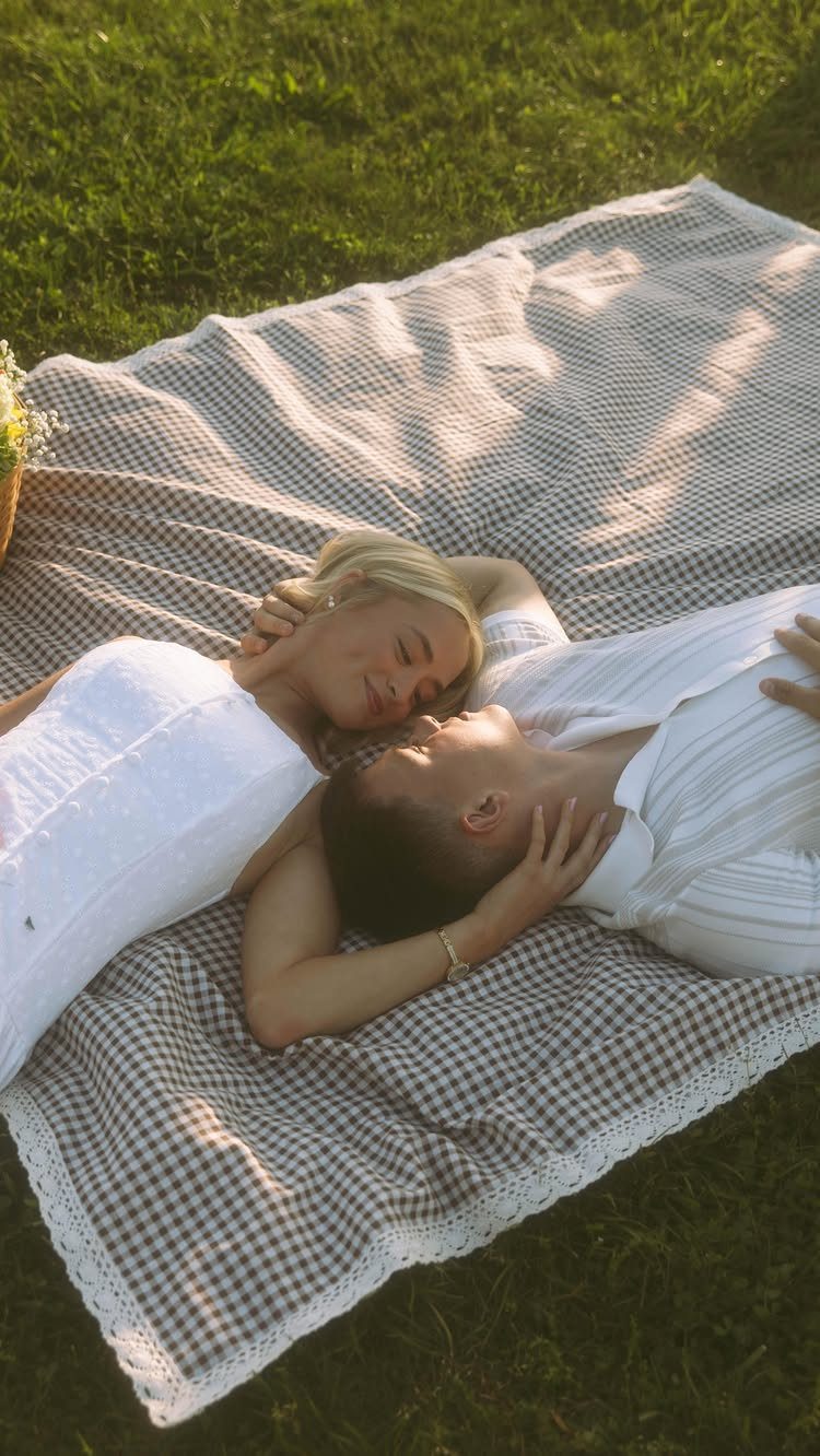 Couple in white clothing lying on a checkered blanket in a sunny park.