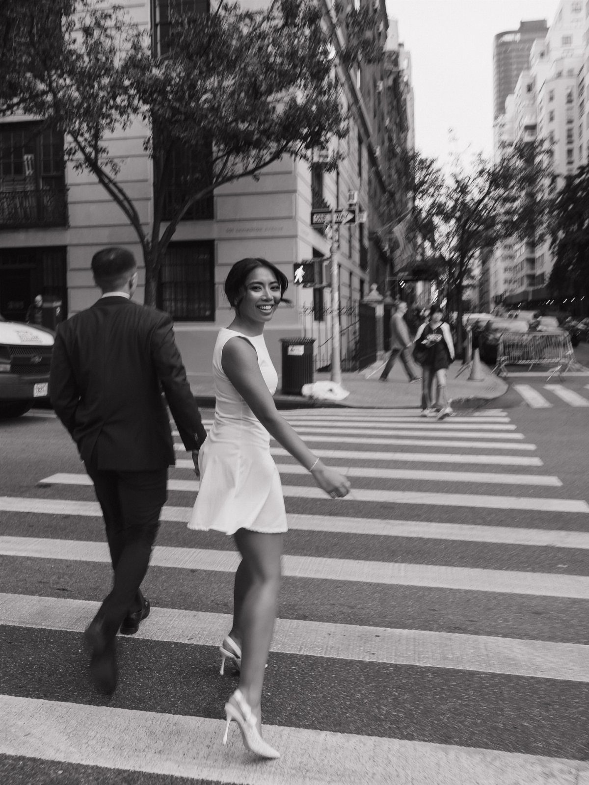Couple walking across a city crosswalk, woman in white dress looking back smiling.