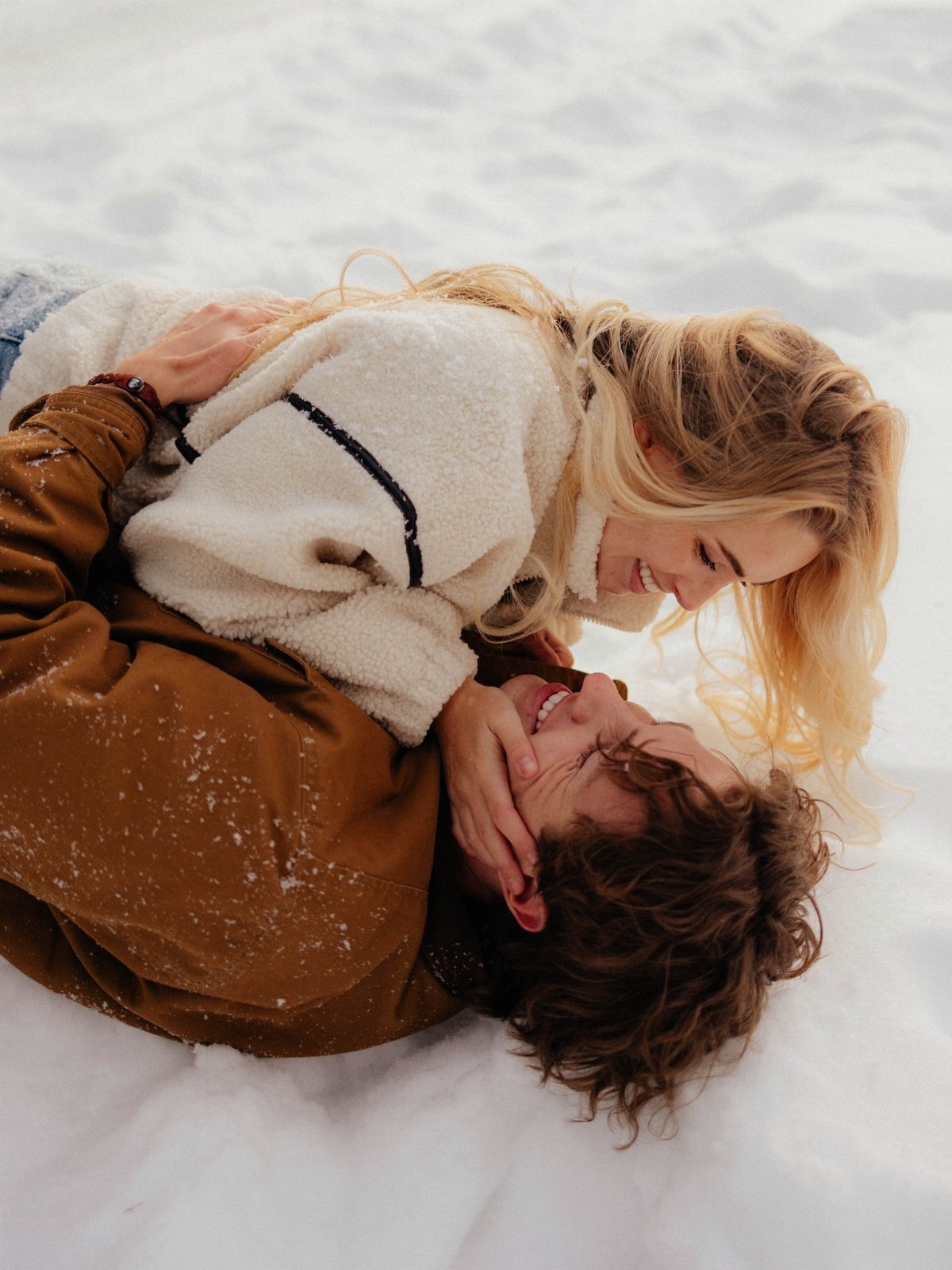 Couple lying in white snow, smiling and looking at each other affectionately.