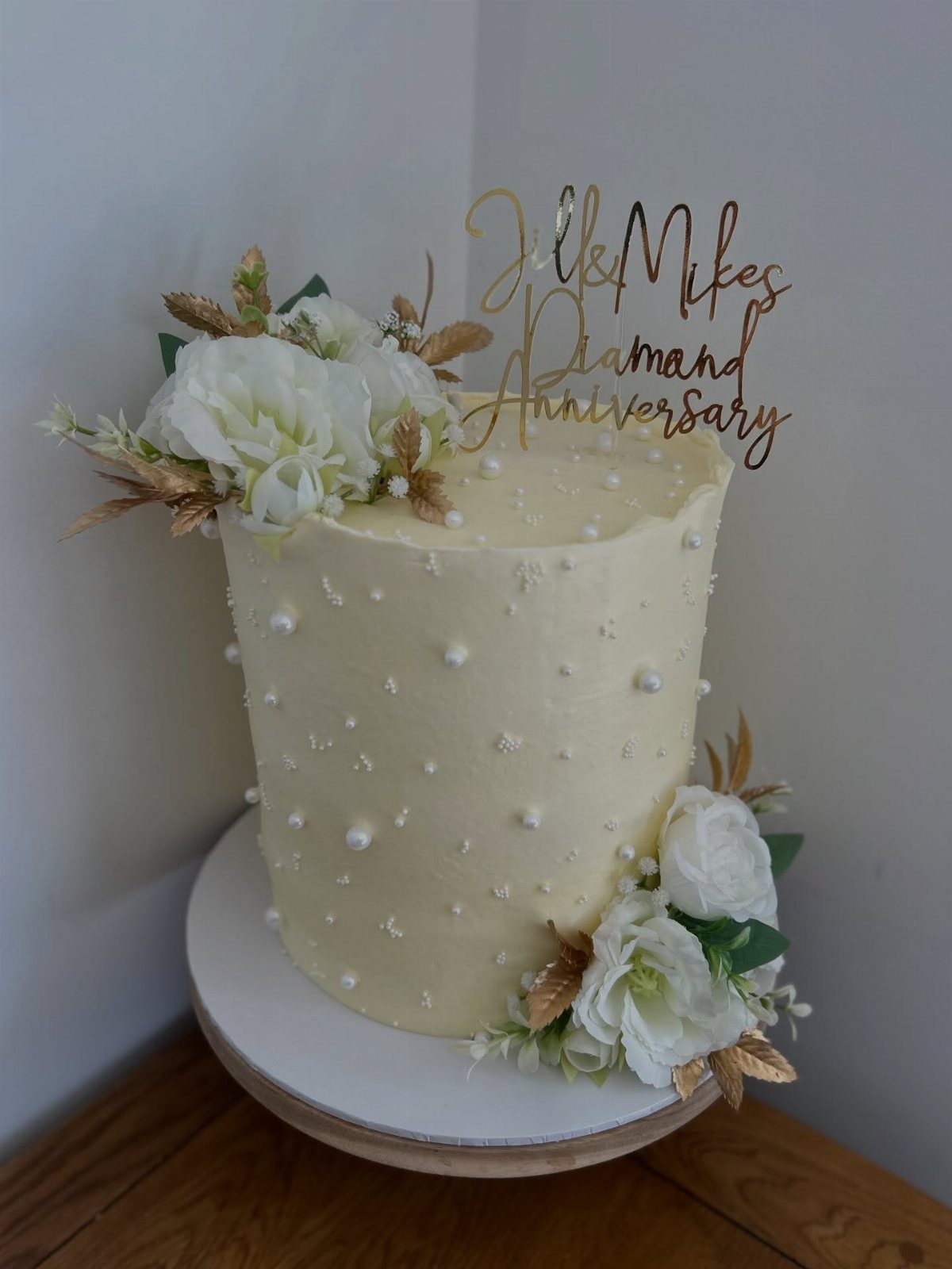 White anniversary cake with edible pearls, white flowers, and gold anniversary topper.
