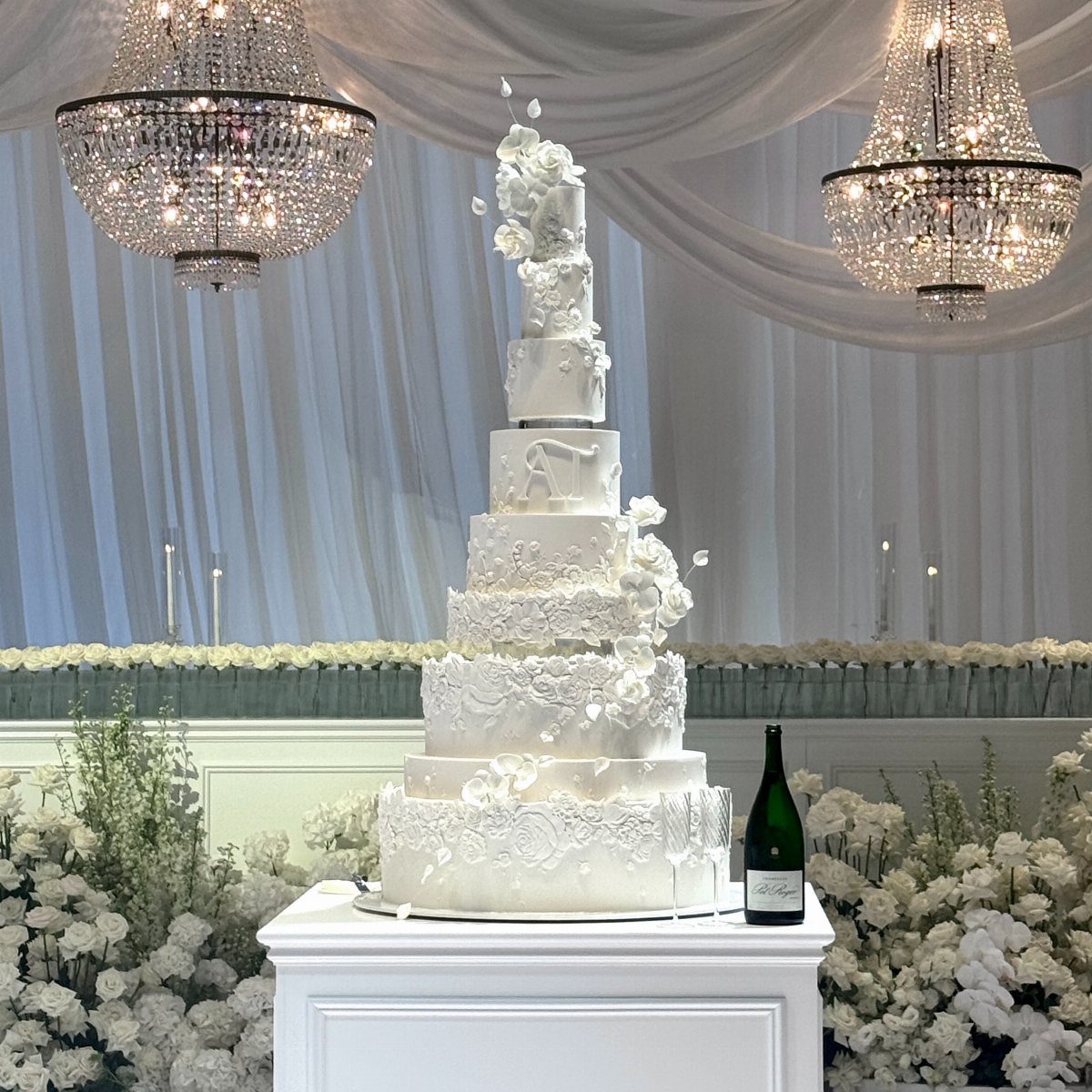 Multi-tiered white fondant cake with lace patterns and floral accents on a pedestal.