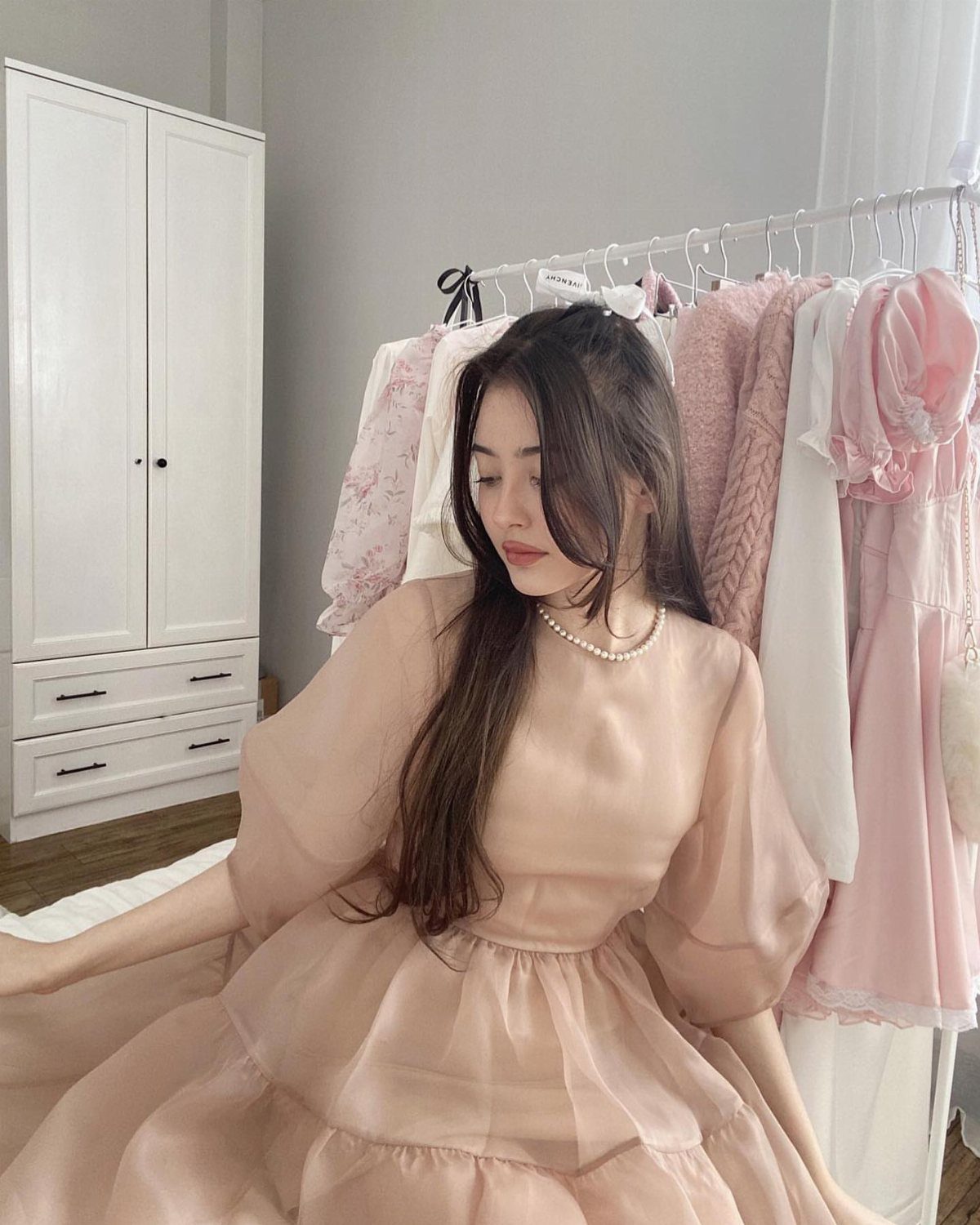Woman in a voluminous beige neutral dress and pearl necklace posing by clothing rack