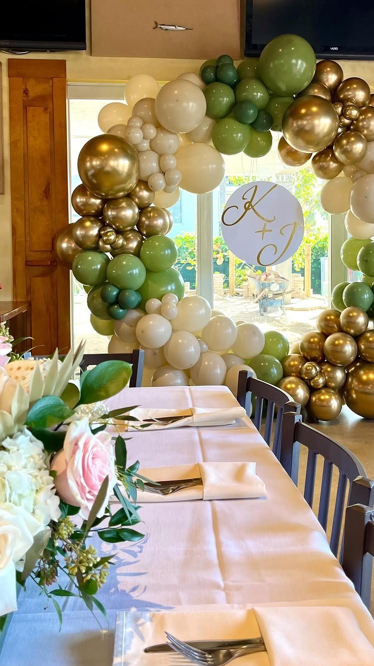 Circular balloon arch in green and gold with initials sign over a decorated table.