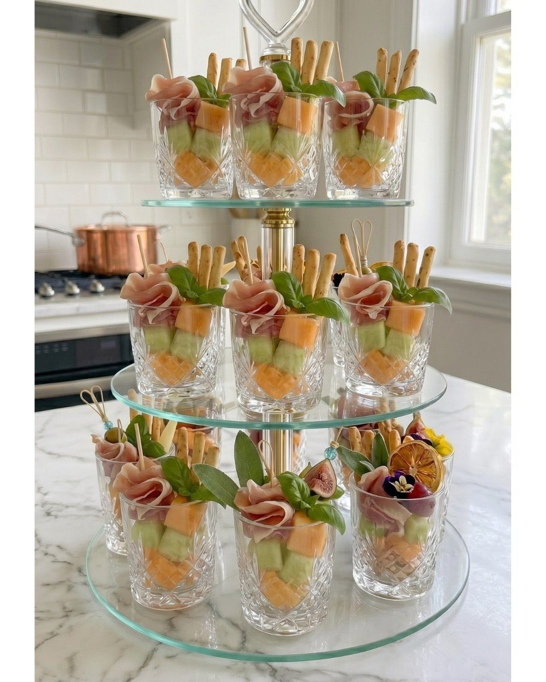 Charcuterie appetizers in small glass cups on a three-tiered glass serving stand.