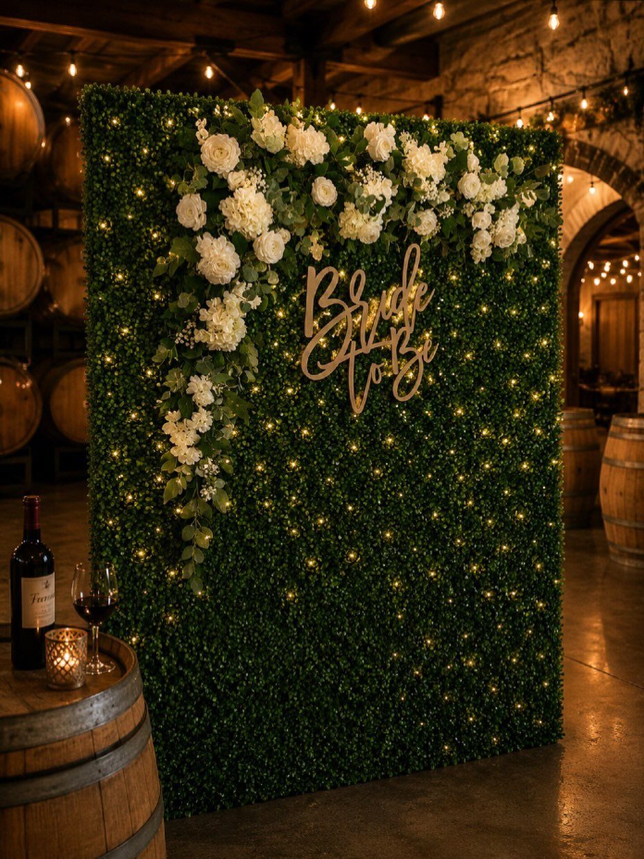 Greenery wall backdrop with white flowers, fairy lights, and a wooden script sign.