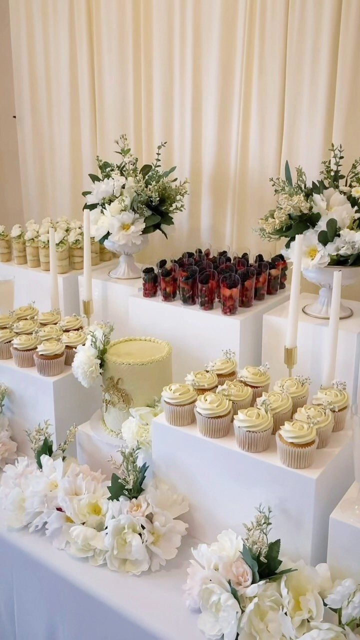 Dessert table with white pedestals, yellow cake, cupcakes, and white floral arrangements.