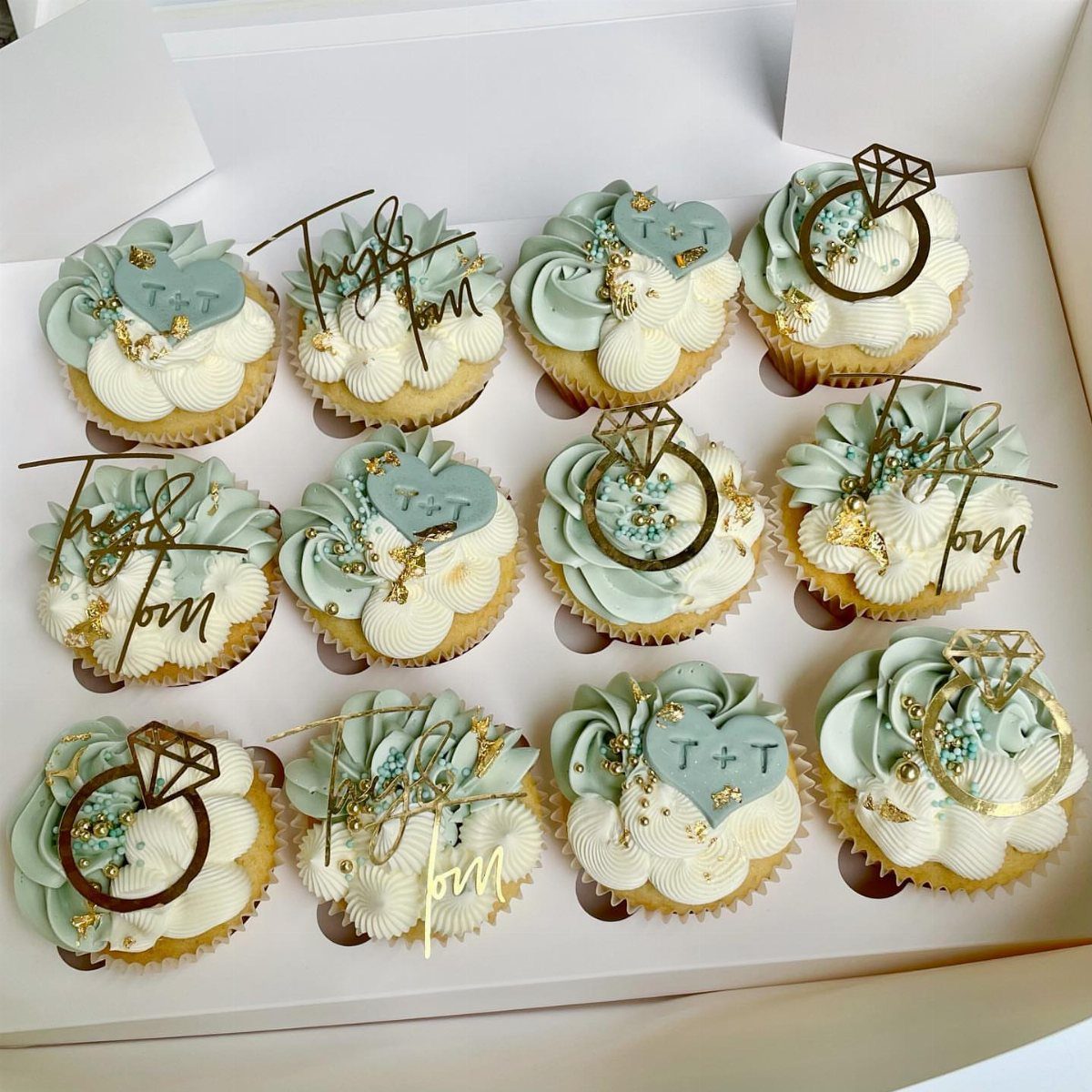 Cupcakes with light blue frosting and gold ring and initial toppers in a box