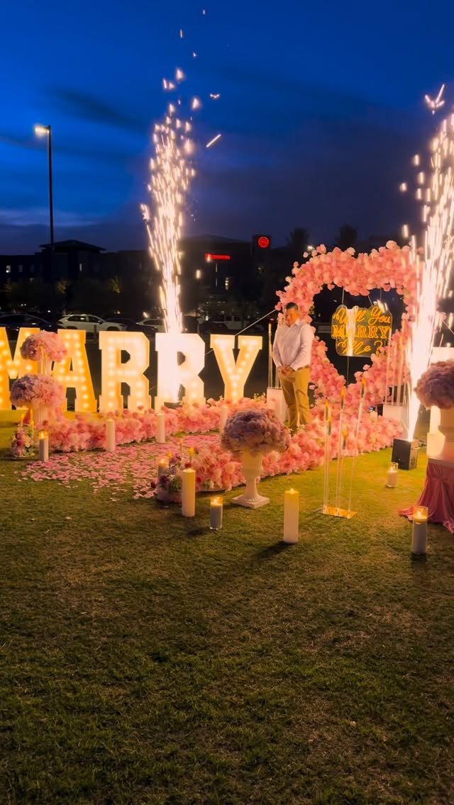 Illuminated marquee letters spelling MARRY surrounded by pink flowers and fireworks at night