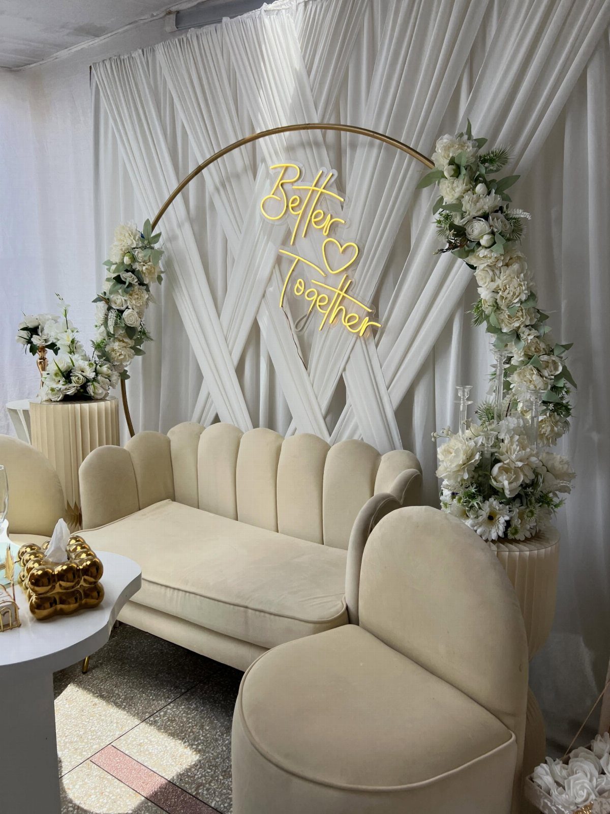 Cream scalloped sofa and chairs under a gold floral arch with neon sign.