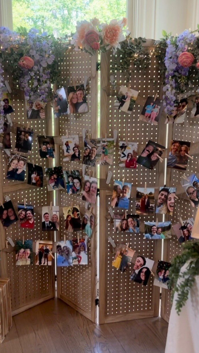 Wooden pegboard screen with clipped photographs, warm lighting, and purple floral arrangements.