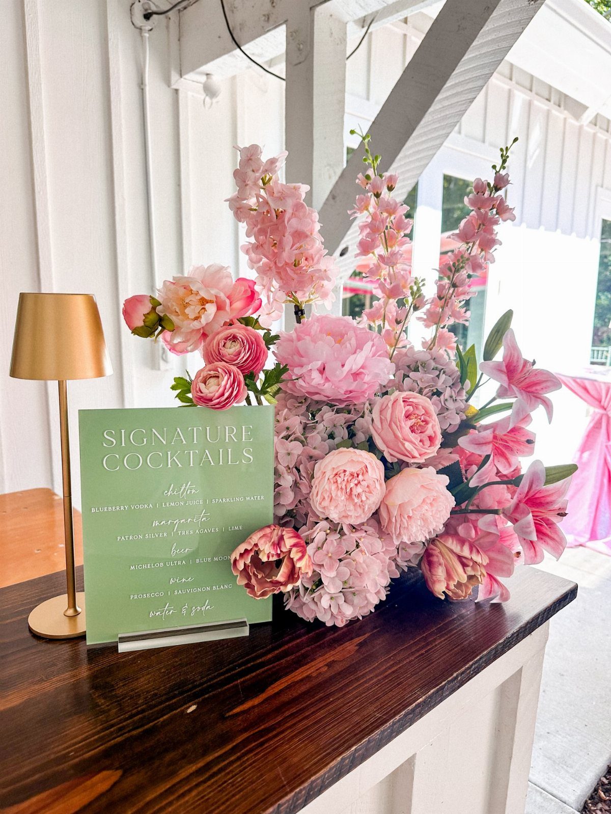 Signature cocktails sign next to a lush arrangement of pink flowers on a bar.