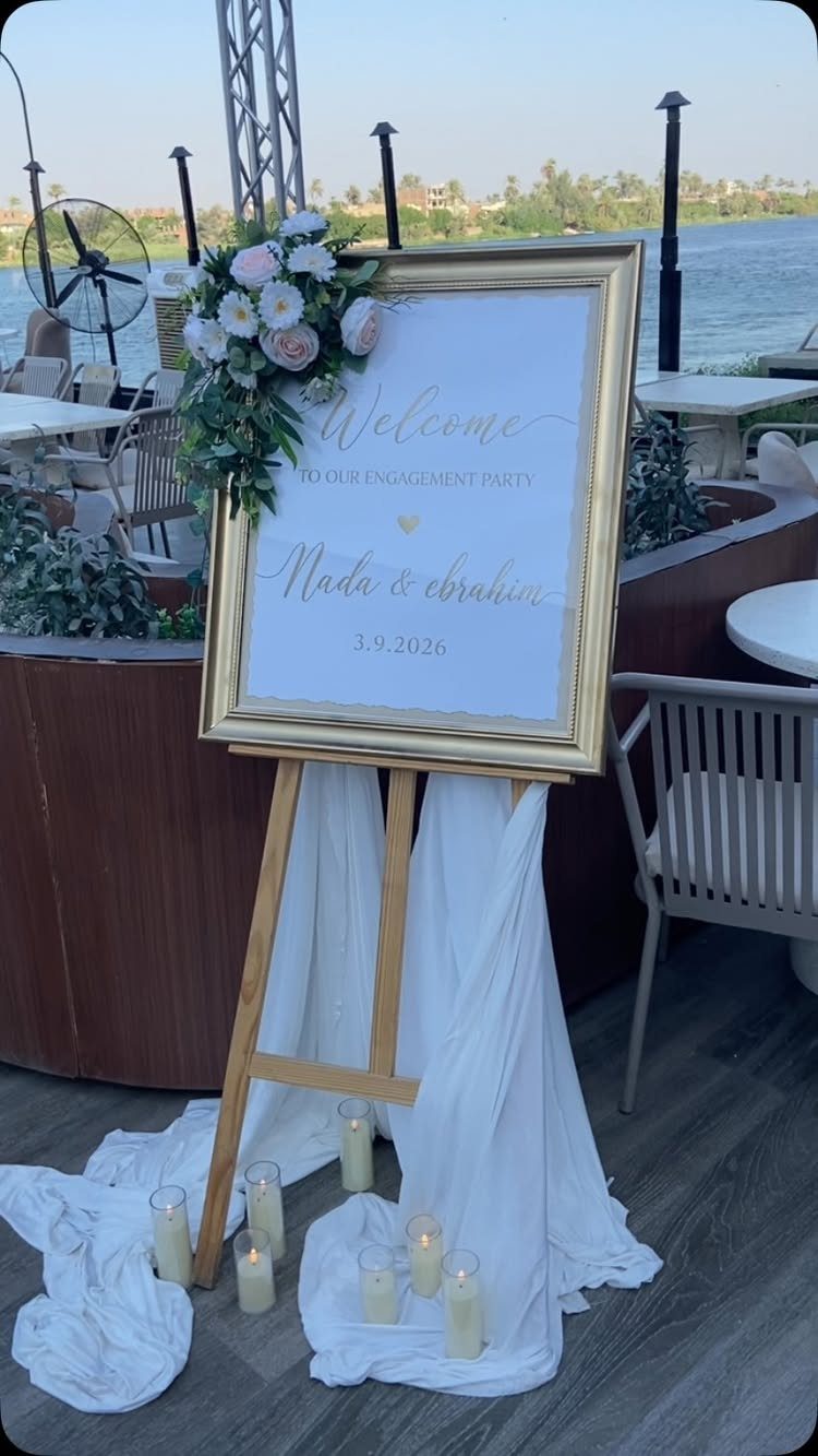 Gold-framed engagement party welcome sign on a draped easel with floral accents and candles.