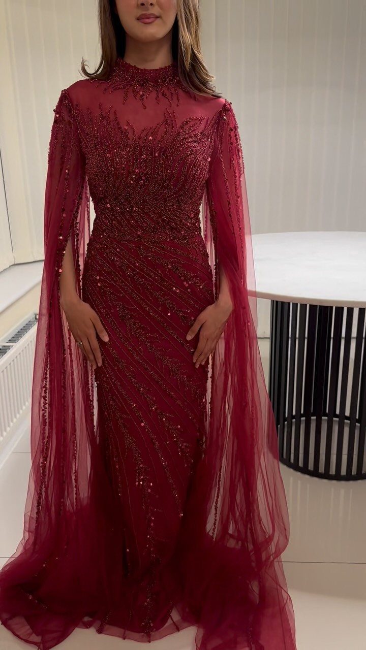 Woman wearing a bold red sequined gown with sheer cape sleeves in a room.
