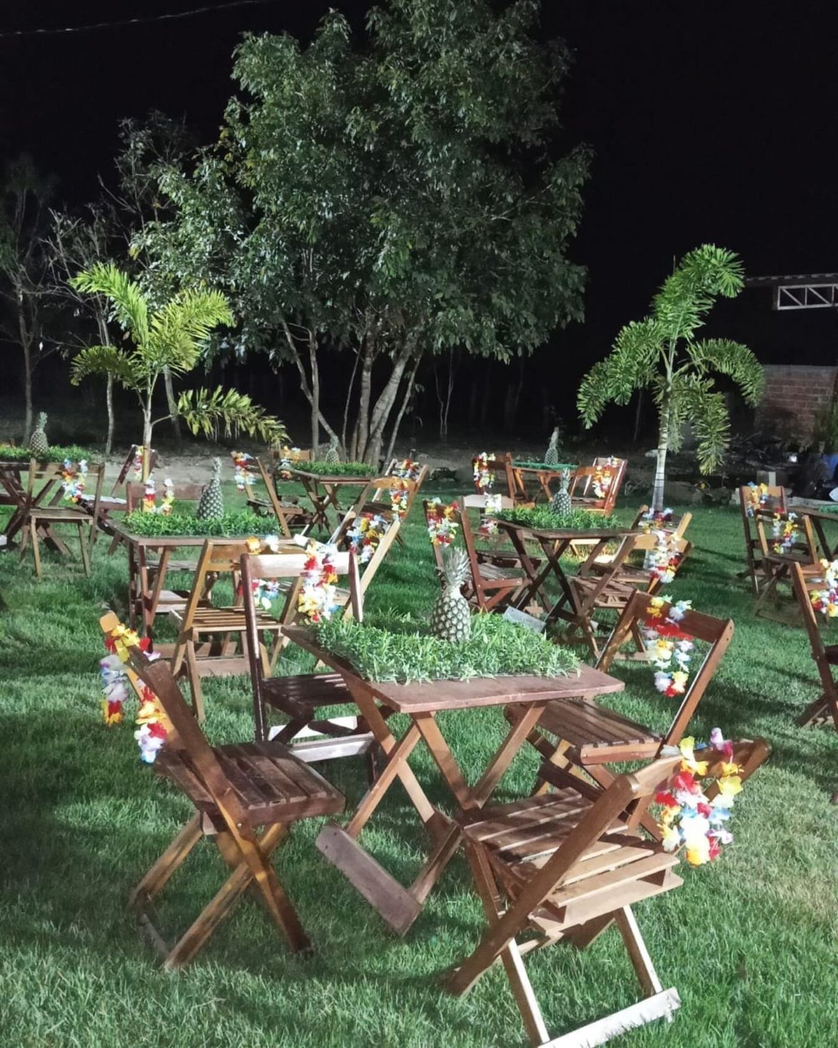 Wooden tables and chairs with pineapples and floral leis in a tropical backyard.