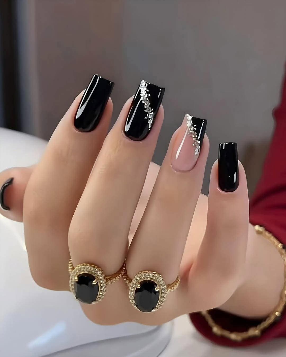 Black and silver rhinestone nail designs on a hand wearing gold rings.