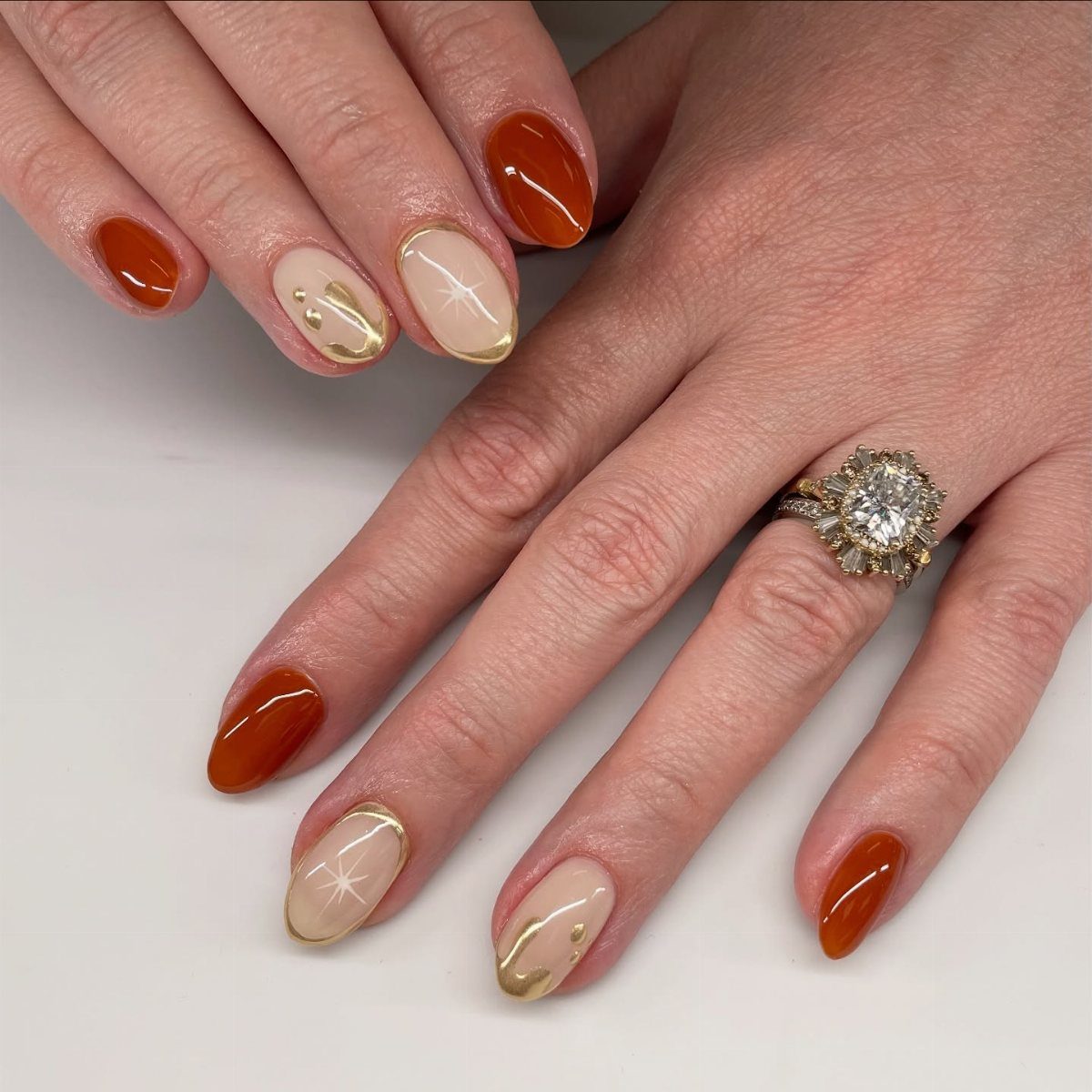 Manicured nails in burnt orange and nude with gold accents and diamond ring.