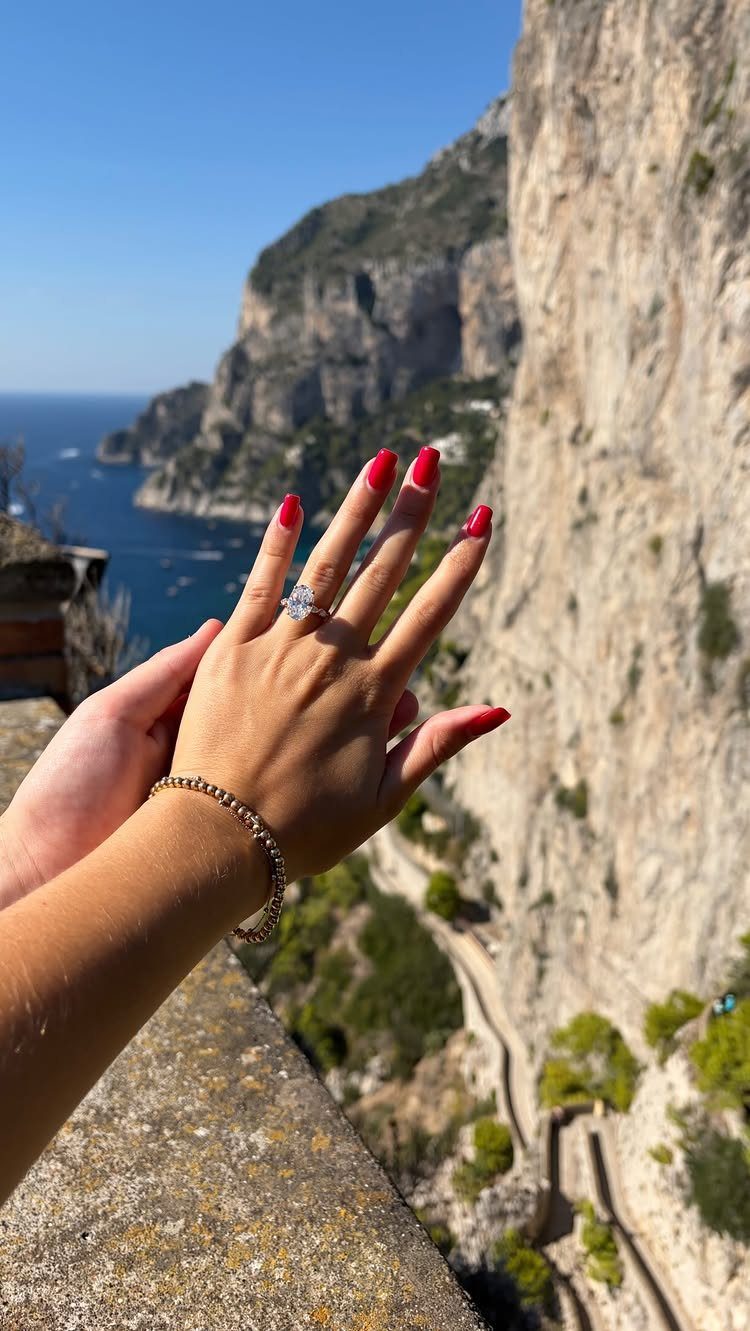 Hand with classic red nails and diamond ring overlooking coastal cliffs and ocean.