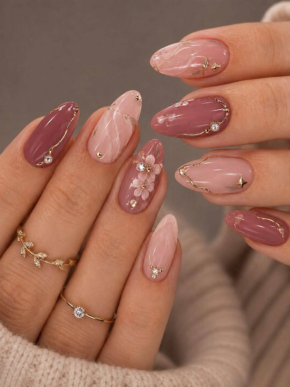 Pink and mauve almond nails with gold swirls, rhinestones, and white floral accents.