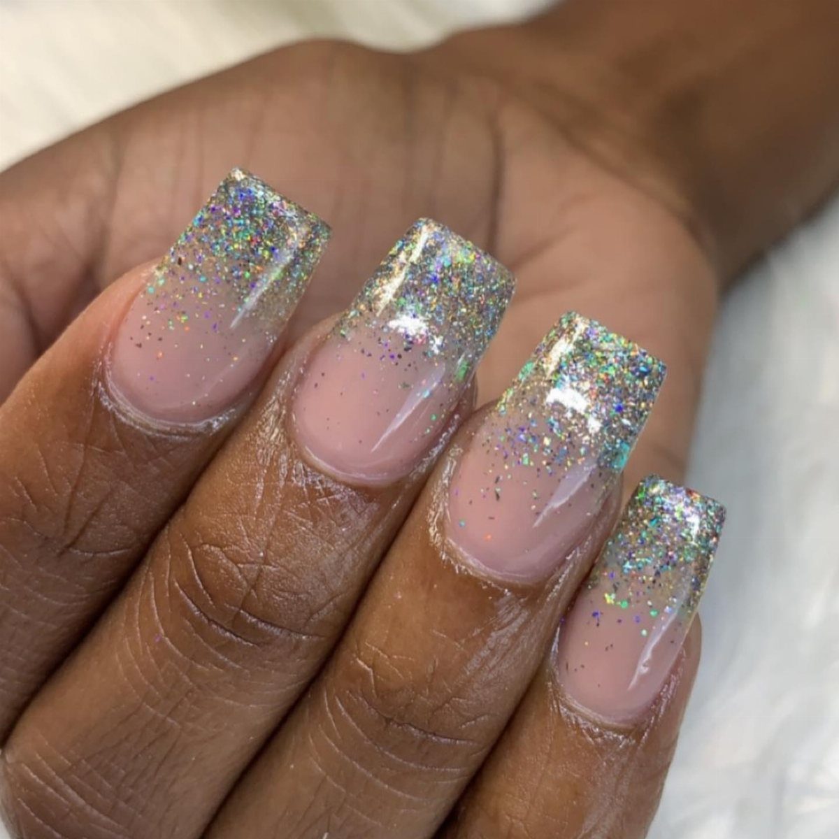 Nude square nails with iridescent holographic glitter tips on a dark-skinned hand.