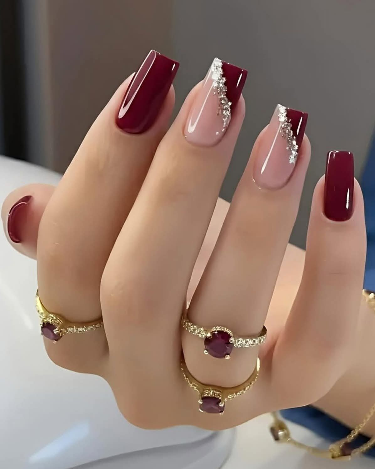 Burgundy and nude coffin nails with rhinestone accents and gold gemstone rings.