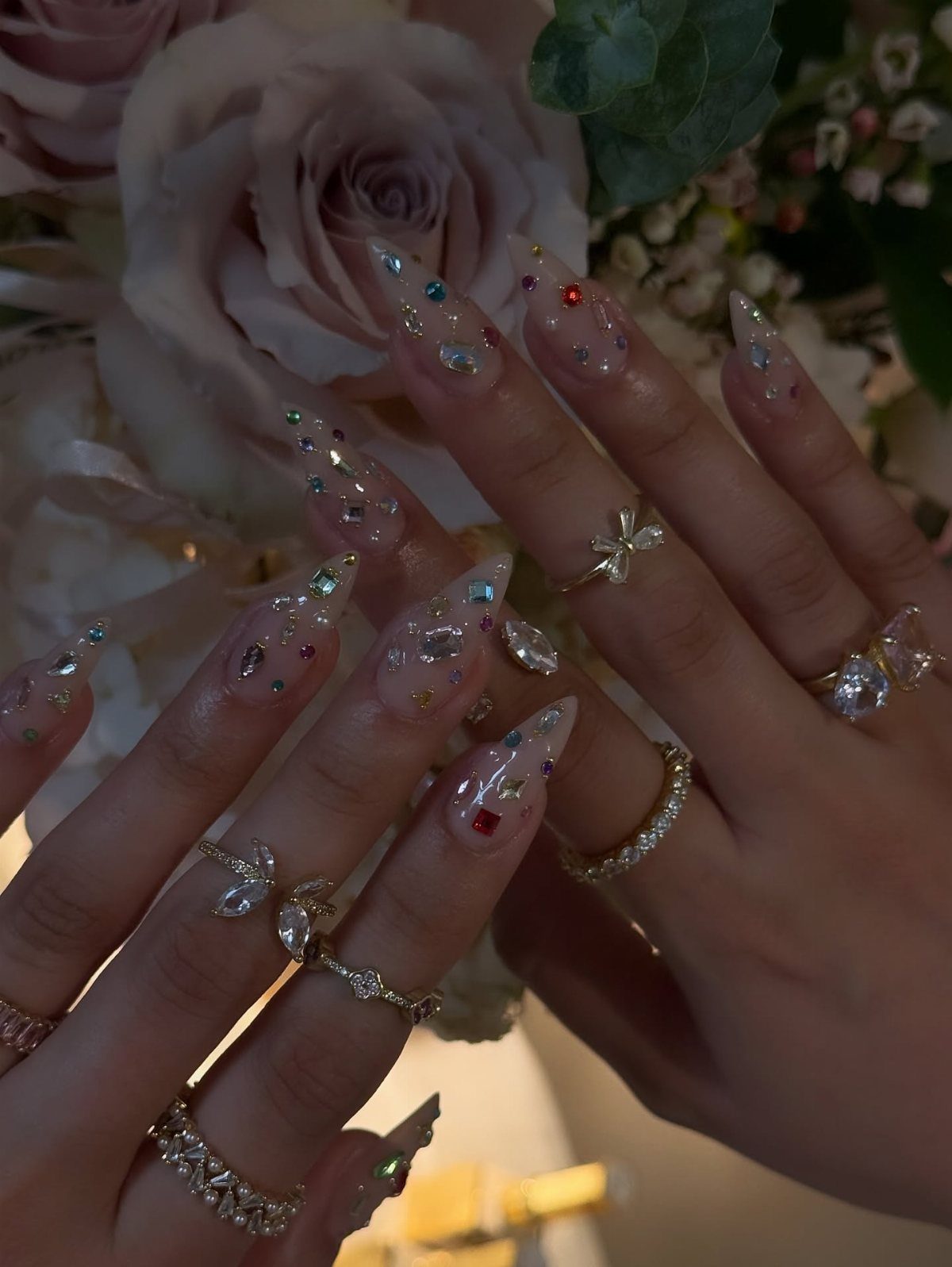 Clear almond nails with colorful rhinestones and gold rings against pink roses