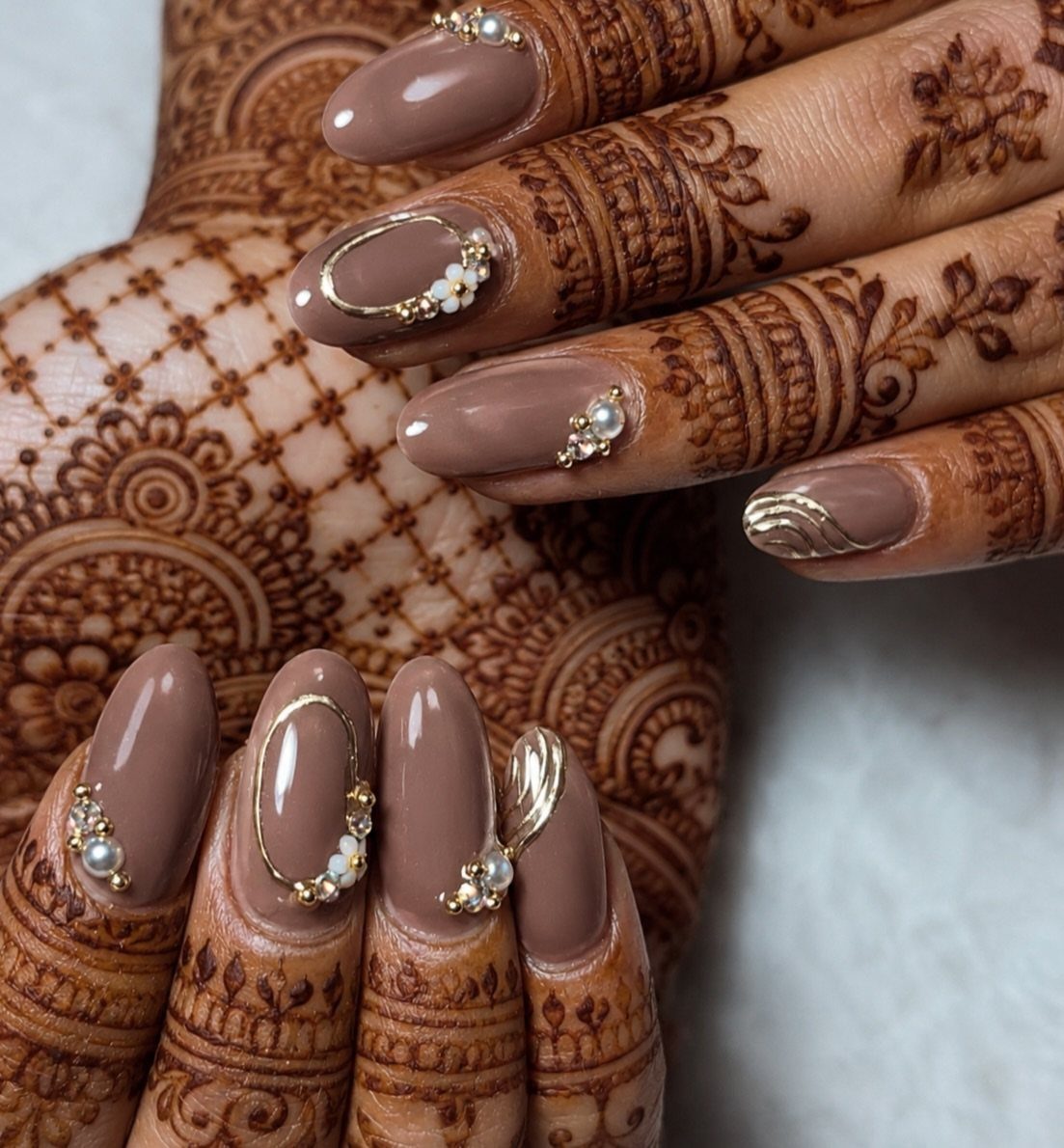 Brown almond nails with 3D gold and pearl accents on henna-decorated hands.
