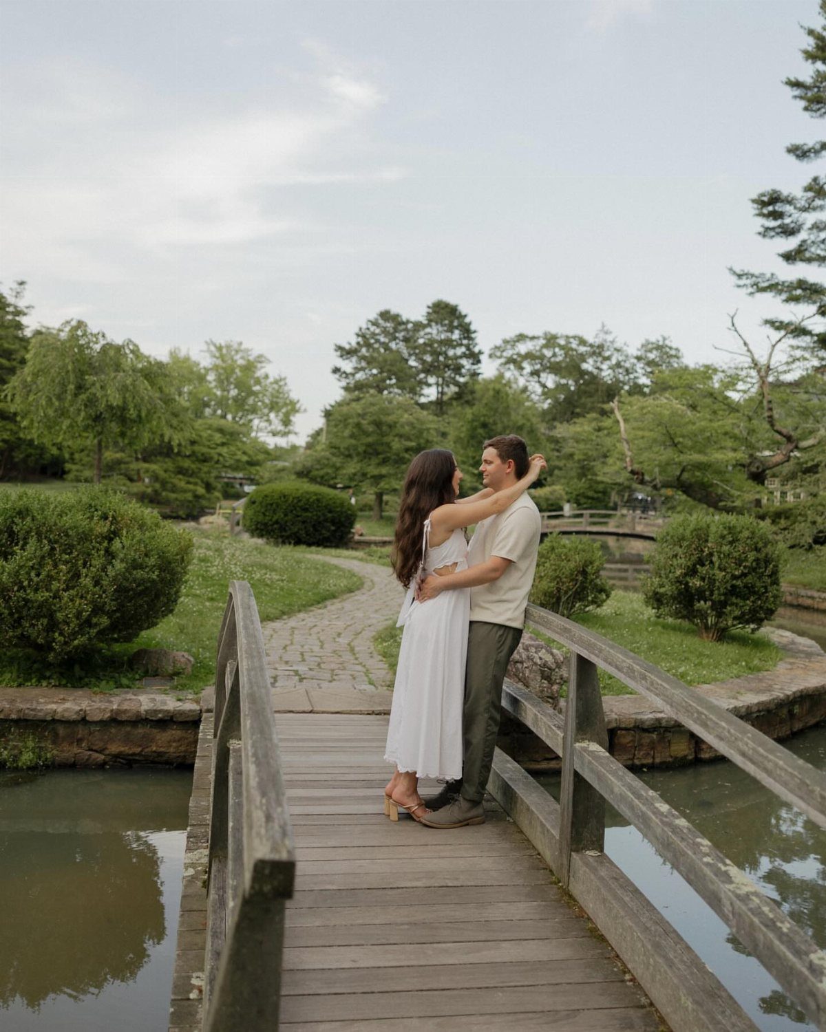 Couple embracing on a wooden bridge in a lush green garden setting.
