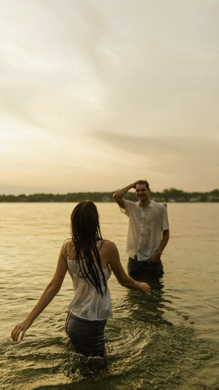 Couple standing in a lake at sunset, facing each other in casual clothing.
