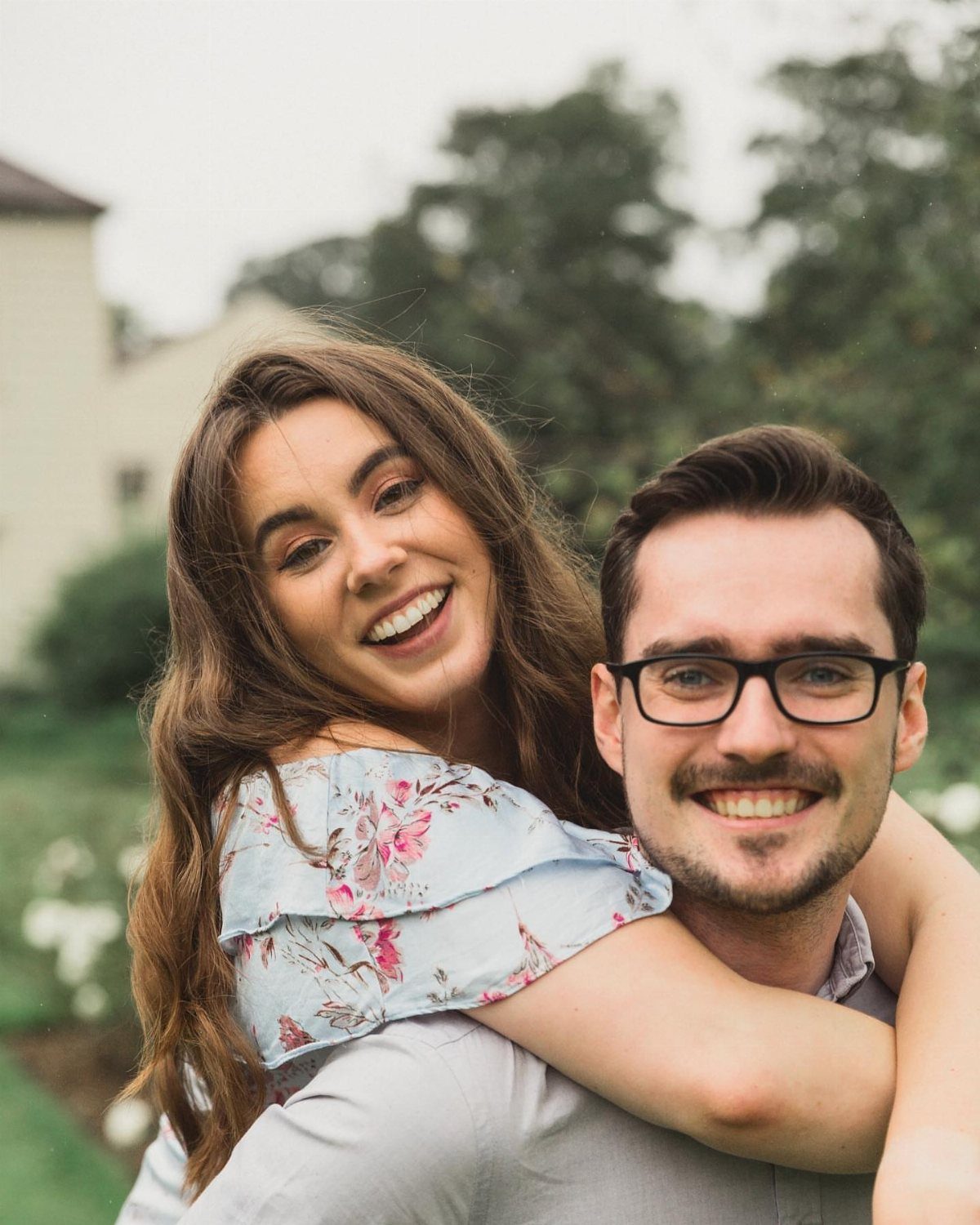 Smiling woman in floral blouse giving a man with glasses a piggyback ride.